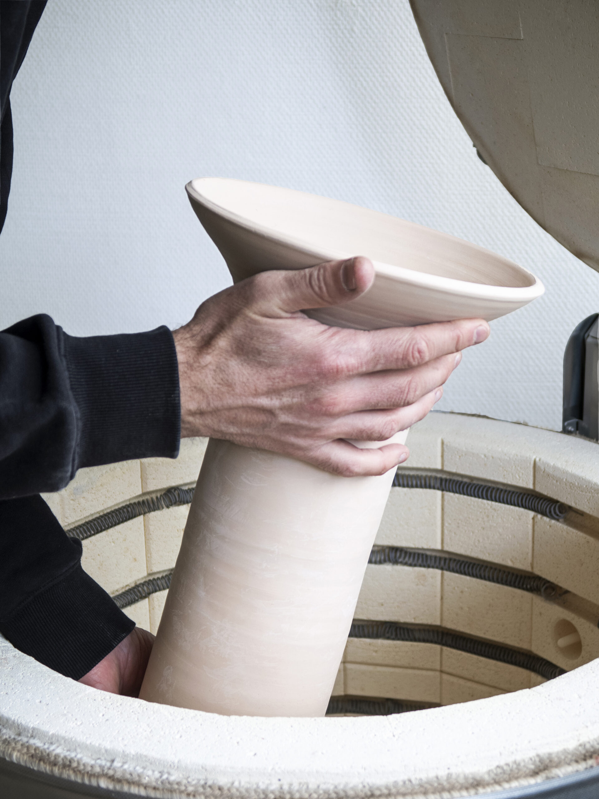Gros plan sur les mains d'un artisan déposant un grand vase en terre brute à l'intérieur d'un four de cuisson de céramique.
