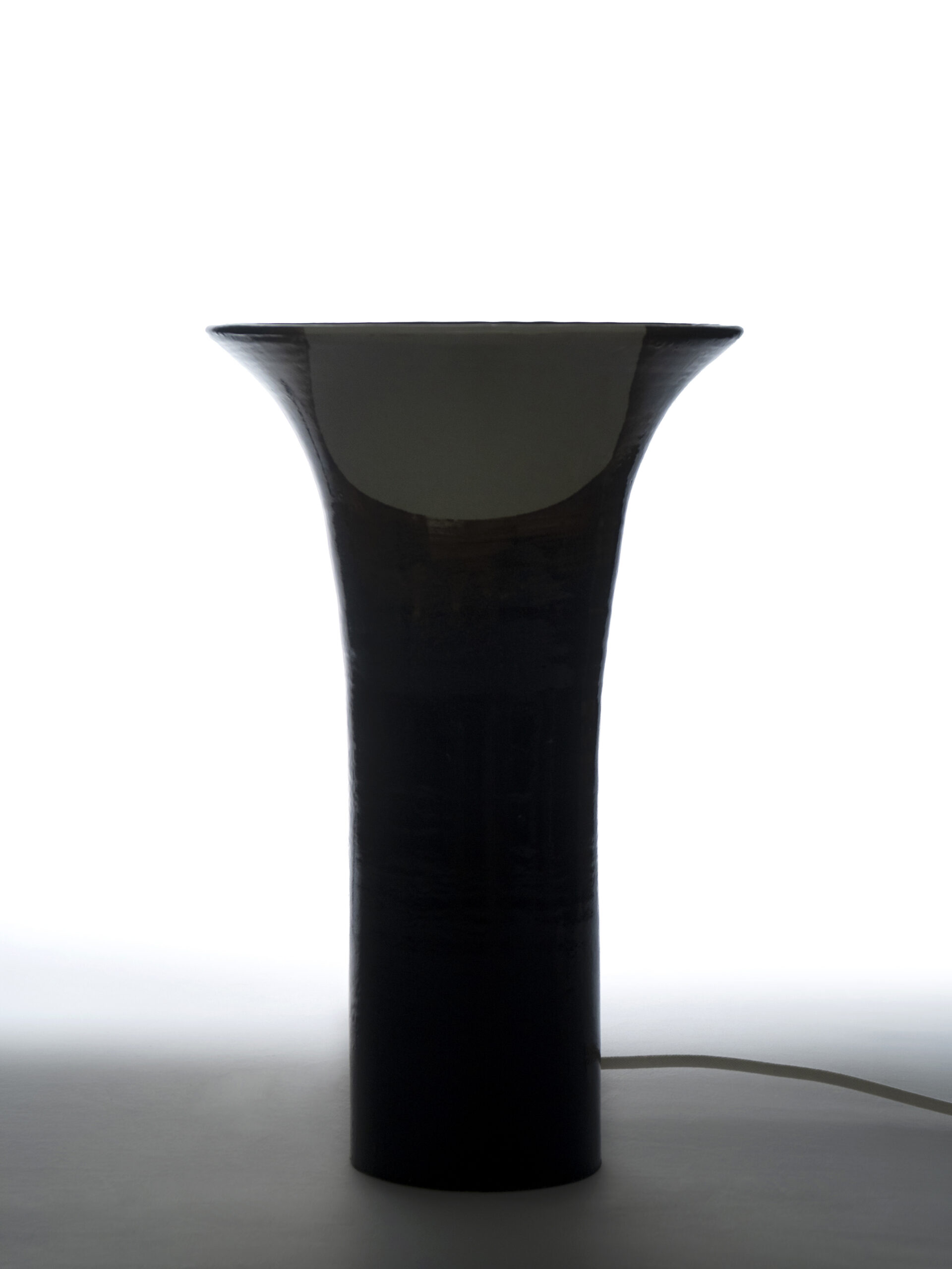 Silhouette rétroéclairée d'un grand  luminaire LUCIEN en céramique noire et blanche avec un col évasé et un câble visible.