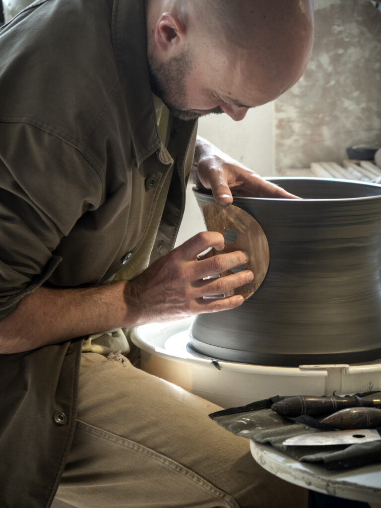 Artisan céramiste à l'atelier façonnant une grande pièce en argile sur un tour de potier en utilisant une estèque en métal.